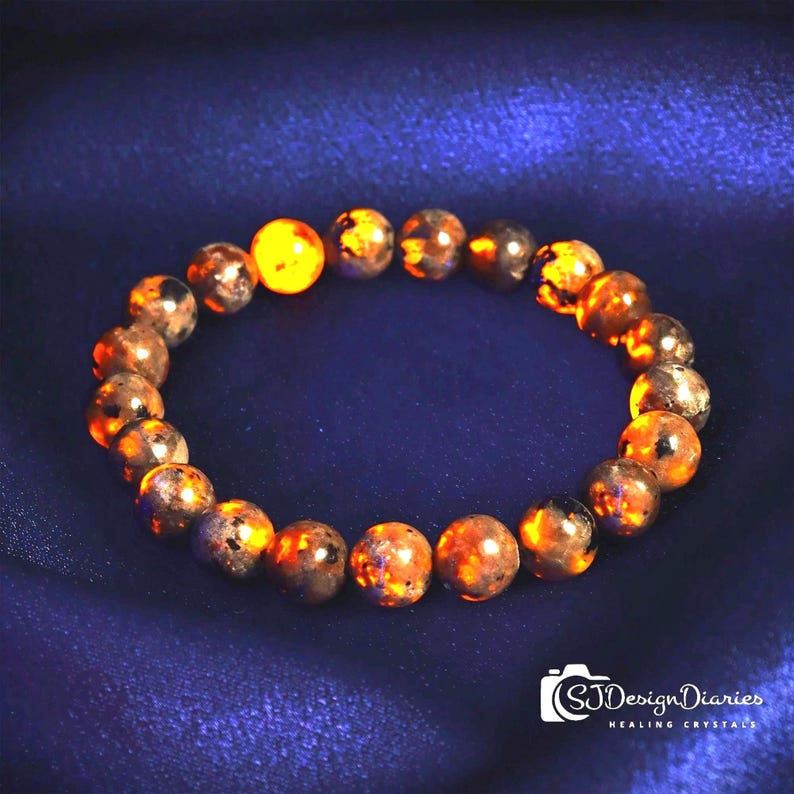 May include: A beaded bracelet featuring round, polished stones in shades of orange, brown, and black. The bracelet is displayed on a dark blue, textured surface. The beads have a speckled appearance, with a mix of colors and textures.
