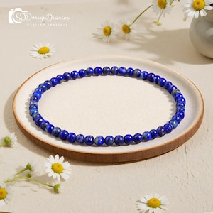 May include: A beaded bracelet made of small, round, deep blue lapis lazuli stones. The bracelet is displayed on a light brown plate, surrounded by white daisies. The text "SJ Design Diaries Healing Crystals" is in the upper left corner.