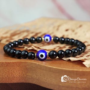 May include: A black beaded bracelet featuring two blue and white evil eye charms. The bracelet is composed of small, round, polished black beads. The evil eye charms are blue with a white iris and black pupil, and are accented with gold-coloured metal.