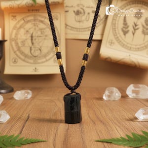 以下が含まれることがあります： 黒トルマリンクリスタルペンダントネックレス、濃い茶色の編み込みコード付き。 ネックレスは金色のアクセントがあり、木製の表面に水晶とヴィンテージの紙を背景に展示されています。 テキスト「SJDesignDiaries Healing Crystals」が表示されています。