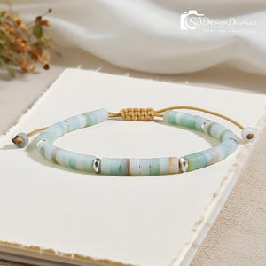 May include: A beaded bracelet featuring light blue, green, and tan disc-shaped beads. The bracelet has a woven beige cord and silver-colored accents. The adjustable closure is a beige cord with a small bead at the end. The bracelet is on a white surface.