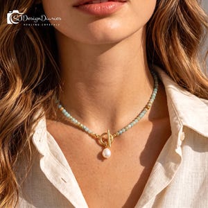 May include: A beaded necklace featuring small, light blue-green gemstone beads and a gold-toned toggle clasp. A single white, round pearl pendant hangs from the centre of the circular toggle ring, creating a delicate and balanced jewellery piece.