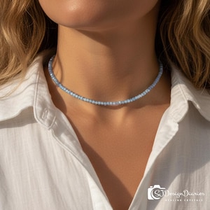 Puede incluir: Un delicado collar de cuentas azul claro descansa sobre el cuello de una persona. El collar está compuesto por pequeñas cuentas redondas, creando un estilo choker sutil y elegante. La persona lleva una camisa blanca abotonada.
