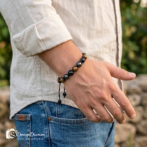 May include: A beaded bracelet featuring alternating round black and golden-brown tiger eye stones. The bracelet is strung on a dark cord with two dangling ends and is worn on a wrist. The design includes smooth, polished spherical beads.