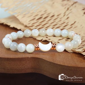 May include: A white beaded bracelet with a crescent moon charm. The bracelet features round, white beads and rose gold accents. The moon charm is white and is flanked by rose gold spacers. The bracelet is displayed on a wooden surface.