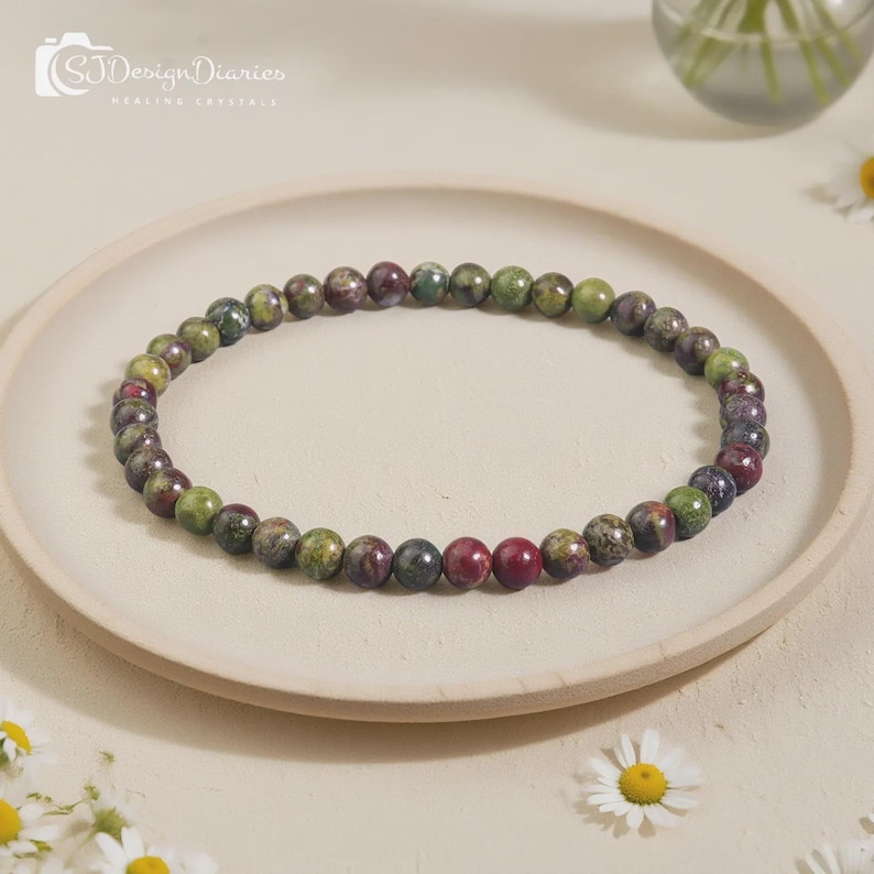 May include: A beaded bracelet composed of small, round, polished stones in variegated shades of forest green, deep burgundy, and dark grey. The beads are strung in a continuous circular loop, displaying natural mineral patterns and textures.