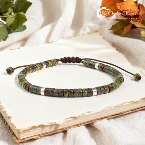 May include: A beaded bracelet featuring green, brown, and silver beads. The bracelet is made with small, square-shaped beads and silver spacers. The bracelet is on a white surface. The bracelet is adjustable.