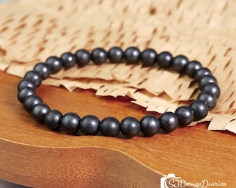 May include: A circular bracelet composed of small, smooth, dark gray spherical beads strung together. The beads have a subtle metallic sheen and are arranged in a continuous loop. The text SJ Design Diaries Healing Crystals is visible.