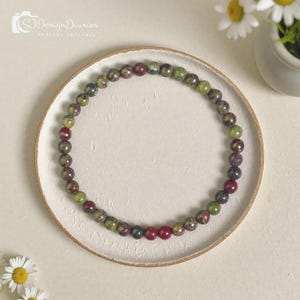 May include: A circular beaded necklace featuring small, polished round stones in variegated shades of forest green, olive, and deep burgundy red. The beads are strung together in a continuous loop, displaying natural color variations and textures.