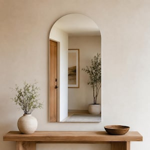 May include: A tall, arched wall mirror with a smooth, frameless edge. The reflective surface shows a room interior with a wooden door, a potted plant, and a framed wall art piece. The mirror is mounted vertically on a plain, light-colored wall.