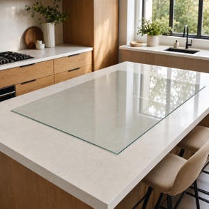 May include: A rectangular, clear glass protective cover resting flat on a light-coloured kitchen island countertop. The glass sheet features smooth, straight edges and a transparent surface that reflects the surrounding kitchen environment.