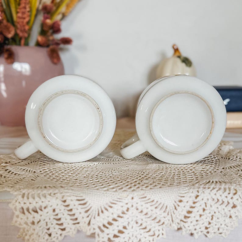 Puede incluir: Dos tazas de cer&aacute;mica blanca colocadas boca abajo para mostrar sus bases circulares. Cada taza presenta un acabado blanco liso y sencillo con una peque&ntilde;a asa en el lateral. Las tazas est&aacute;n situadas una al lado de la otra sobre un tapete de encaje.