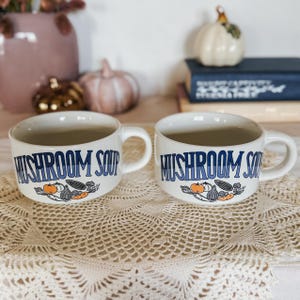 Puede incluir: Dos tazas de sopa de cer&aacute;mica de color crema con el texto MUSHROOM SOUP en letras azules. Debajo del texto, cada taza muestra una ilustraci&oacute;n gr&aacute;fica de varias verduras, incluyendo champi&ntilde;ones, zanahorias y cebollas en naranja y negro.