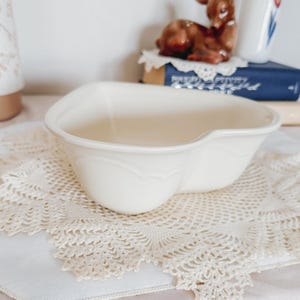 May include: A cream-colored, heart-shaped ceramic bowl with a decorative embossed design. The bowl is sitting on a cream-colored, lace doily. Other decorative items are in the background.