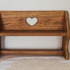 Puede incluir: Un estante de pared de madera de color marrón medio que presenta una tabla horizontal con un recorte en forma de corazón en el centro del panel posterior. La unidad tiene dos soportes laterales con bordes curvos y una barra inferior horizontal.