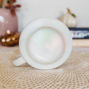 Puede incluir: Una taza de cer&aacute;mica blanca vista desde arriba, que muestra una abertura circular con un borde liso y brillante y un asa que se extiende hacia la izquierda. La base interior de la taza es visible, con un acabado iridiscente y nacarado.