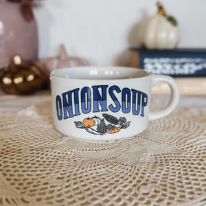Puede incluir: Una taza de sopa de cer&aacute;mica blanca con el texto ONION SOUP en letras azules. Debajo del texto hay una ilustraci&oacute;n gr&aacute;fica de varias verduras, incluyendo un tomate, cebolla, ma&iacute;z y zanahoria en colores naranja, negro y blanco.