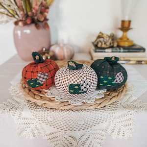 May include: Three fabric pumpkins featuring distinct patterns: one in red plaid, one in a brown and white checkered print, and one in dark green plaid. Each pumpkin is accented with a small heart-shaped patch and a green fabric stem on top.