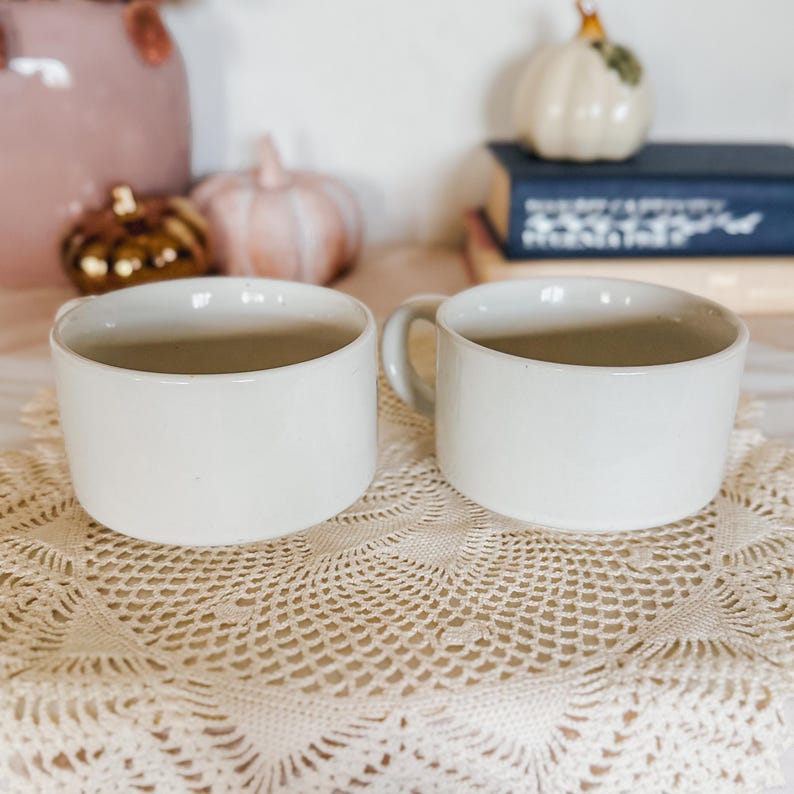 Puede incluir: Dos tazas de cer&aacute;mica de color blanco roto a juego con una forma cil&iacute;ndrica lisa y un asa sencilla en el lateral de cada una. Las tazas tienen un acabado limpio de color s&oacute;lido y est&aacute;n colocadas una al lado de la otra.