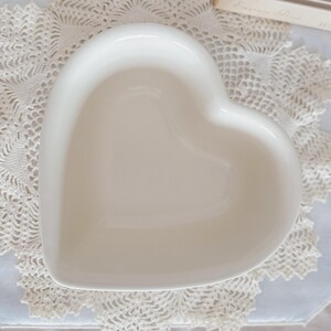May include: A white, heart-shaped ceramic dish. The dish has a smooth, glossy finish and is empty. The heart-shaped dish is sitting on a white lace doily. The dish is likely used for serving or display.
