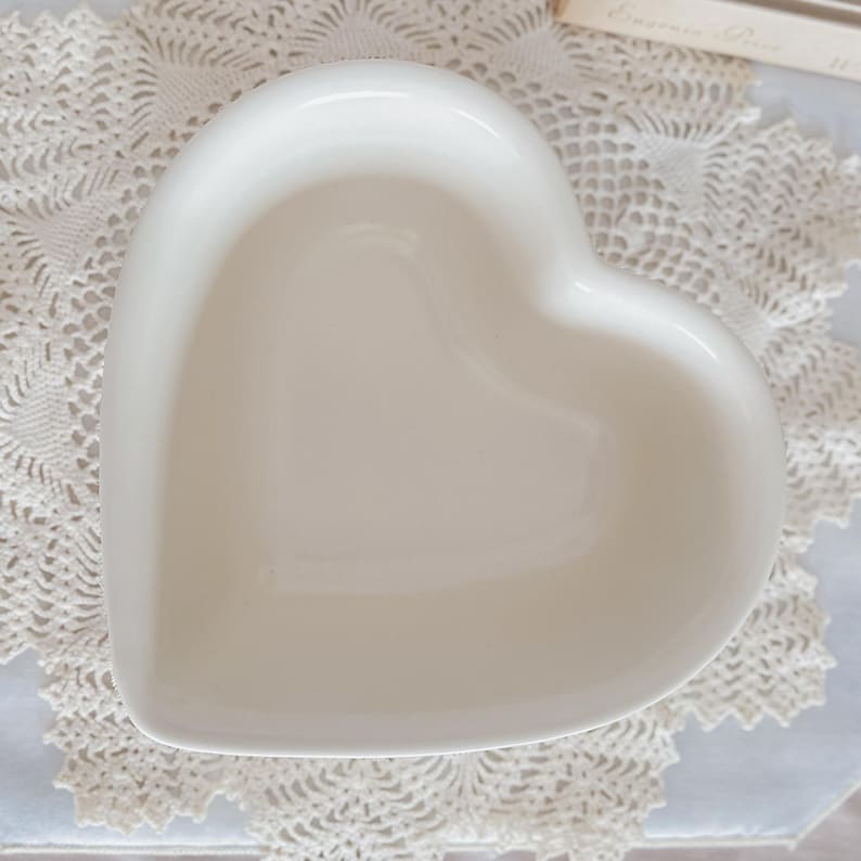 May include: A white, heart-shaped ceramic dish. The dish has a smooth, glossy finish and is empty. The heart-shaped dish is sitting on a white lace doily. The dish is likely used for serving or display.