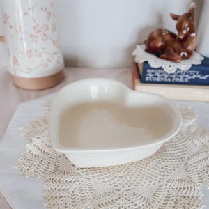 May include: A heart-shaped, off-white ceramic dish. The dish sits on a cream-colored, lace doily. A decorative ceramic deer figurine and books are in the background. A floral-patterned ceramic pitcher is also visible.