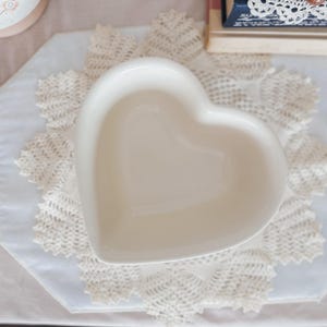 May include: A heart-shaped, off-white ceramic dish. The dish is placed on a white lace doily. The background includes a stack of books and a decorative item. The dish is empty and has a smooth, glossy finish.