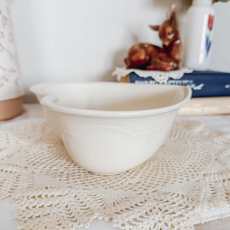 May include: A cream-colored ceramic mixing bowl with a scalloped design. The bowl sits on a cream-colored lace doily. In the background, a small brown deer figurine and a white vase with a red and blue design are visible.
