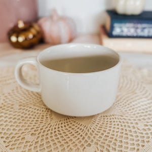 Puede incluir: Una taza de cer&aacute;mica ancha de color blanco roto con un acabado mate liso y un asa redondeada. La taza presenta una forma cil&iacute;ndrica baja con una base plana, dise&ntilde;ada para contener bebidas calientes. La superficie es lisa, sin patrones ni texto.