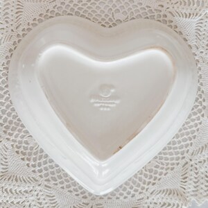 May include: A white heart-shaped ceramic dish. The dish has a smooth, glossy surface and a slightly raised edge. The bottom of the dish has a stamp that reads "Hallmark USA". The dish is placed on a white lace doily.