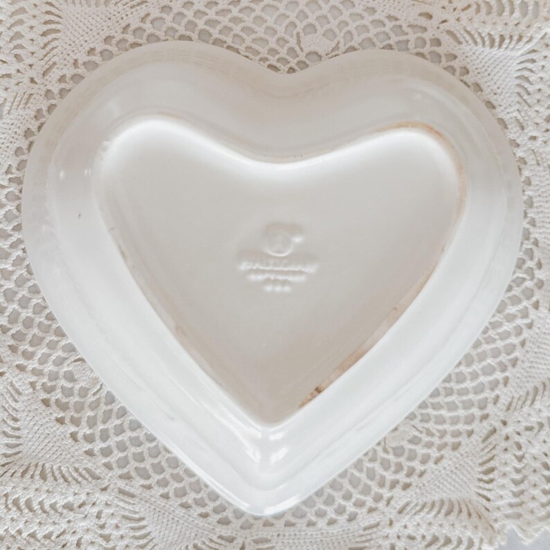 May include: A white heart-shaped ceramic dish. The dish has a smooth, glossy surface and a slightly raised edge. The bottom of the dish has a stamp that reads "Hallmark USA". The dish is placed on a white lace doily.