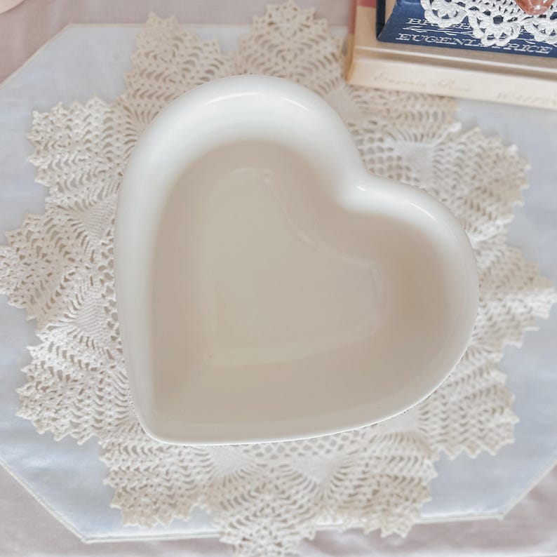 May include: A heart-shaped, off-white ceramic dish. The dish is placed on a white and cream-colored lace doily. The dish is empty and has a smooth, glossy finish. The background includes books and other decorative items.