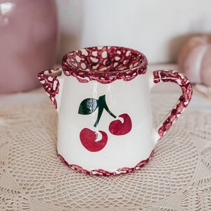 Puede incluir: Una jarrita de leche de cerámica blanca con un motivo de cerezas rojas pintado a mano y hojas verdes. El borde y el asa están decorados con un patrón festoneado pintado con esponja en rojo y blanco, dando una textura.