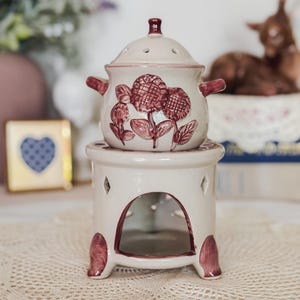 May include: A cream-colored ceramic oil warmer featuring a removable top bowl with a dark red sunflower relief design and two side handles. The base is a cylindrical stand with a large arched opening for a tea light and decorative cutouts.