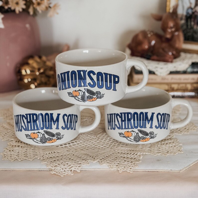 Puede incluir: Tres cuencos de sopa de cer&aacute;mica color blanco roto con asas. Un cuenco presenta el texto azul ONION SOUP, mientras que dos cuencos presentan el texto azul MUSHROOM SOUP. Cada cuenco est&aacute; decorado con una peque&ntilde;a ilustraci&oacute;n de verduras en naranja y gris.