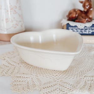 May include: Off-white heart-shaped ceramic bowl with a scalloped edge. The bowl sits on a cream-colored lace doily. A decorative deer figurine and books are in the background.