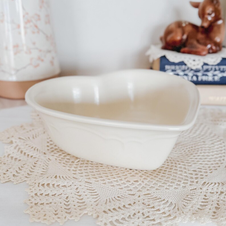 May include: Off-white heart-shaped ceramic bowl with a scalloped edge. The bowl sits on a cream-colored lace doily. A decorative deer figurine and books are in the background.