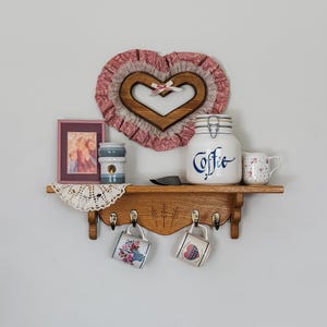 May include: A wooden shelf with a heart-shaped decoration, a framed photo, and a coffee jar. Two decorative mugs hang below. The shelf is adorned with a doily and a small creamer. The coffee jar has the word "Coffee" written on it.