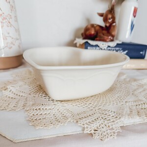 May include: Cream-colored heart-shaped ceramic bowl with scalloped detailing. The bowl sits on a cream-colored lace doily. A brown ceramic deer figurine and a floral-decorated container are in the background.