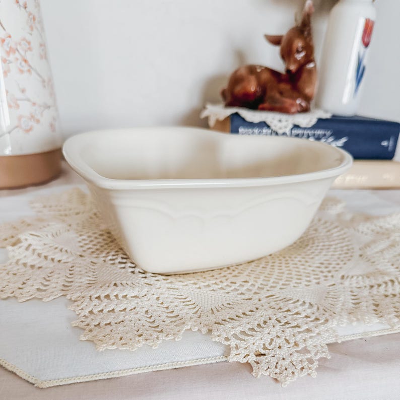May include: Cream-colored heart-shaped ceramic bowl with scalloped detailing. The bowl sits on a cream-colored lace doily. A brown ceramic deer figurine and a floral-decorated container are in the background.