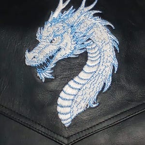 May include: Close-up of a black leather jacket pocket featuring a white and blue embroidered dragon. The dragon's design is detailed, with visible scales and a curved neck. The embroidery adds texture to the jacket's surface.