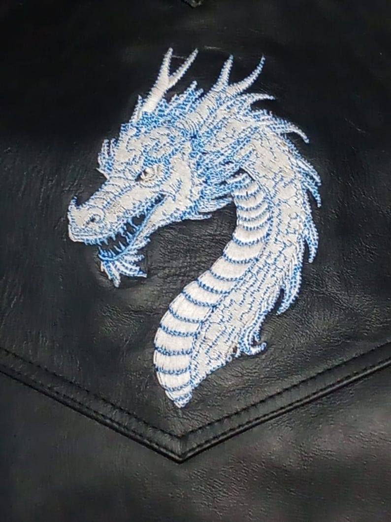 May include: Close-up of a black leather jacket pocket featuring a white and blue embroidered dragon. The dragon's design is detailed, with visible scales and a curved neck. The embroidery adds texture to the jacket's surface.