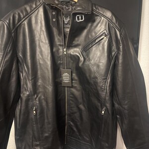 May include: Black leather jacket with a zip-up front, a small chest pocket, and a collar. The jacket has a tag attached. The jacket is hanging on a wooden hanger.