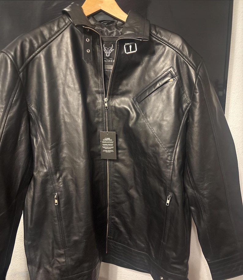 May include: Black leather jacket with a zip-up front, a small chest pocket, and a collar. The jacket has a tag attached. The jacket is hanging on a wooden hanger.