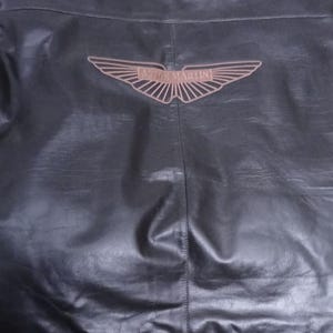 May include: Black leather jacket featuring the Aston Martin logo on the back. The logo is a brown wing design with the brand name. The jacket has a classic design.