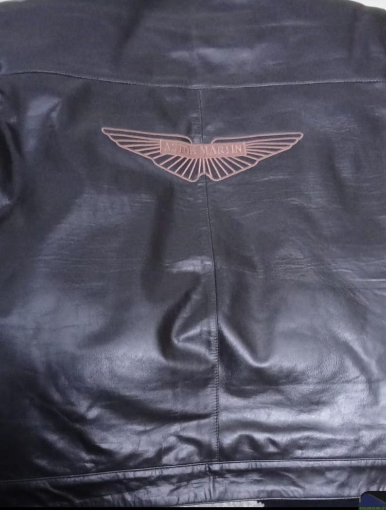 May include: Black leather jacket featuring the Aston Martin logo on the back. The logo is a brown wing design with the brand name. The jacket has a classic design.
