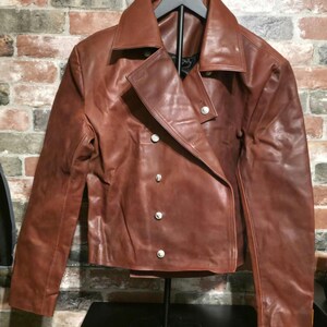 May include: A brown leather jacket with a wide collar and silver buttons. The jacket has a double-breasted design and is displayed on a black stand. The leather appears distressed, giving it a vintage look.