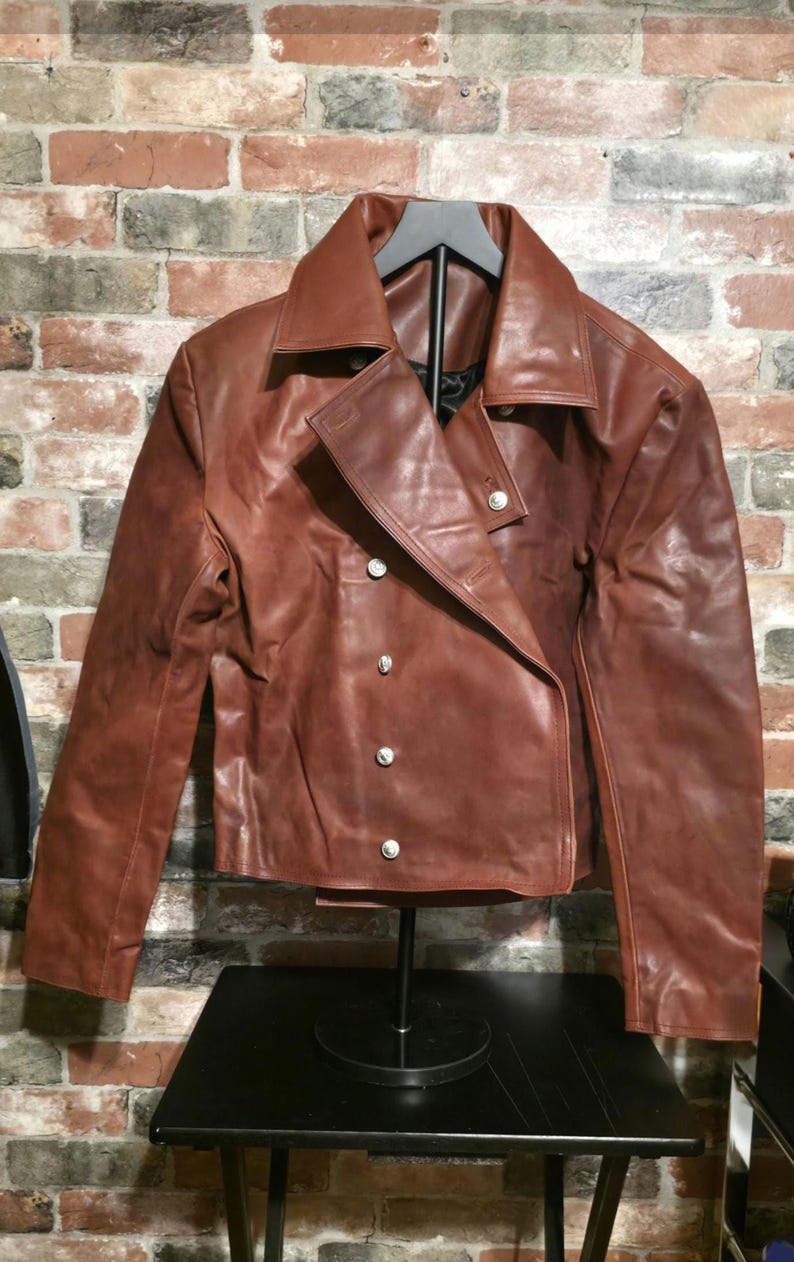 May include: A brown leather jacket with a wide collar and silver buttons. The jacket has a double-breasted design and is displayed on a black stand. The leather appears distressed, giving it a vintage look.