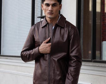 Può includere: Una giacca in pelle marrone scuro con colletto classico, chiusura frontale con zip integrale e maniche lunghe con polsini. La giacca presenta una finitura liscia e lucida ed è indossata sopra una camicia bianca con jeans blu.