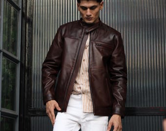 May include: A dark brown leather jacket with a mandarin collar and a front zip fastening, worn over a beige and white vertical striped button-down shirt paired with plain white trousers. The jacket features a smooth finish and long sleeves.